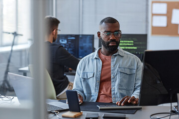 Wall Mural - Portrait of black man using computer while programming mobile software in office behind glass wall