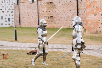 two knights in ancient metal armor are standing at the stone wall of the castle fighting with swords