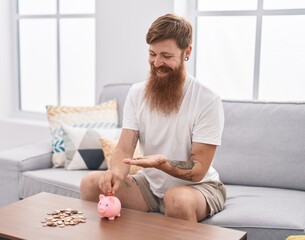 Canvas Print - Young redhead man inserting coin on piggy bank at home