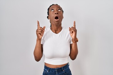 Wall Mural - Beautiful black woman standing over isolated background amazed and surprised looking up and pointing with fingers and raised arms.