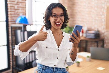 Sticker - Young hispanic woman working at the office using smartphone smiling happy and positive, thumb up doing excellent and approval sign