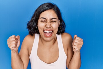 Sticker - Young hispanic woman standing over blue background excited for success with arms raised and eyes closed celebrating victory smiling. winner concept.