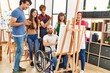 Group of young paint students smiling happy and looking draw of disabled partner sitting on wheelchair at art studio.