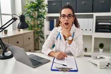 Sticker - Young hispanic woman wearing doctor uniform and stethoscope looking fascinated with disbelief, surprise and amazed expression with hands on chin