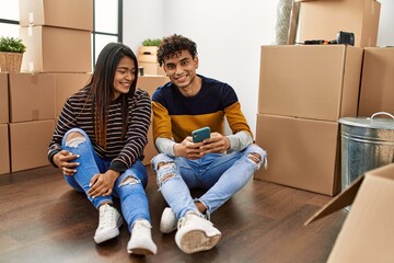 Poster - Young latin couple using smartphone sitting on the floor at new home.