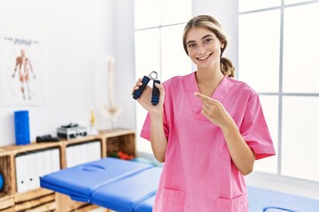 Poster - Young blonde woman working at pain recovery clinic holding hand strengthener smiling happy pointing with hand and finger