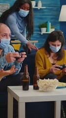 Sticker - Vertical video: Concentrated multiracial friends playing video games with joystick controller in living room wearing face mask to prevent spreading coronavirus. Diverse people having fun, laughing at