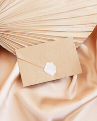 Wall Mural - Sealed envelope on beige satin fabric and dried palm leaf close up, greeting or wedding mockup