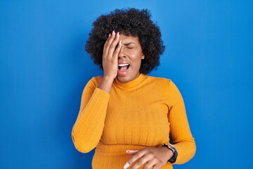 Poster - Black woman with curly hair standing over blue background yawning tired covering half face, eye and mouth with hand. face hurts in pain.