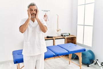 Wall Mural - Middle age hispanic therapist man working at pain recovery clinic with hand on head, headache because stress. suffering migraine.