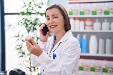 Sticker - Middle age woman pharmacist holding pills bottle talking on smartphone at pharmacy