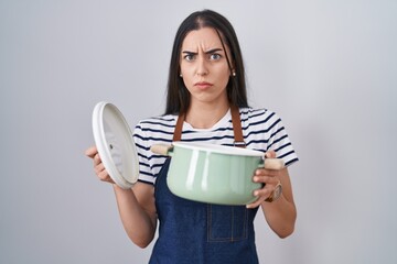 Sticker - Young brunette woman wearing apron holding cooking pot skeptic and nervous, frowning upset because of problem. negative person.