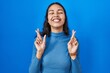 © Krakenimages.com - Young brazilian woman standing over blue isolated background gesturing finger crossed smiling with hope and eyes closed. luck and superstitious concept.