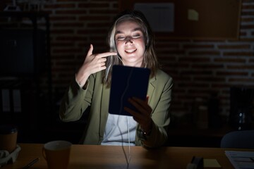 Poster - Blonde caucasian woman working at the office at night smiling cheerful showing and pointing with fingers teeth and mouth. dental health concept.