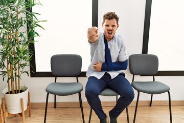 Poster - Handsome young man sitting at doctor waiting room with arm injury pointing displeased and frustrated to the camera, angry and furious with you