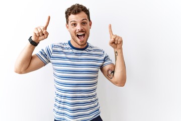 Sticker - Handsome young man standing over isolated background smiling amazed and surprised and pointing up with fingers and raised arms.