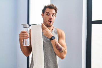 Wall Mural - Young hispanic man wearing sportswear drinking water surprised pointing with finger to the side, open mouth amazed expression.