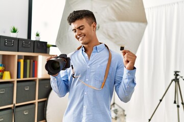 Wall Mural - Young hispanic man photographer holding professional camera and memory card at photography studio