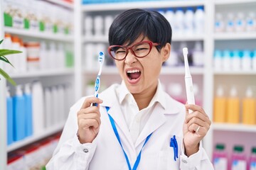 Sticker - Young asian woman with short hair doing toothbrush comparative at pharmacy angry and mad screaming frustrated and furious, shouting with anger. rage and aggressive concept.