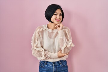 Wall Mural - Young asian woman standing over pink background looking confident at the camera smiling with crossed arms and hand raised on chin. thinking positive.