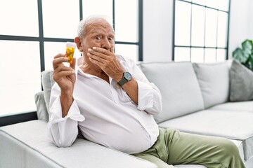 Wall Mural - Senior man holding pills laughing and embarrassed giggle covering mouth with hands, gossip and scandal concept