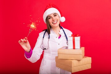 A female doctor in a Santa Claus hat holds boxes with gifts and a sparkler on a red background. Doctor's congratulations to patients...