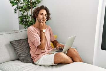 Canvas Print - Young hispanic man sitting on the sofa at home using laptop looking confident at the camera smiling with crossed arms and hand raised on chin. thinking positive.