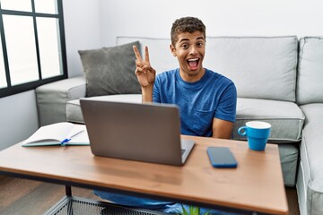 Wall Mural - Young handsome hispanic man using laptop sitting on the floor smiling with happy face winking at the camera doing victory sign. number two.