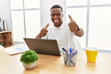 Canvas Print - Young african man working at the office using computer laptop approving doing positive gesture with hand, thumbs up smiling and happy for success. winner gesture.