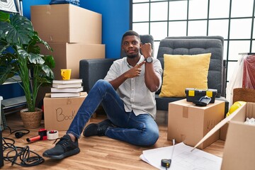 Wall Mural - African american man sitting on the floor at new home in hurry pointing to watch time, impatience, looking at the camera with relaxed expression