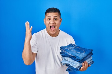 Canvas Print - Hispanic young man holding stack of folded jeans celebrating victory with happy smile and winner expression with raised hands