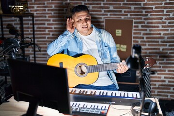 Sticker - Hispanic young man playing classic guitar at music studio smiling with hand over ear listening an hearing to rumor or gossip. deafness concept.