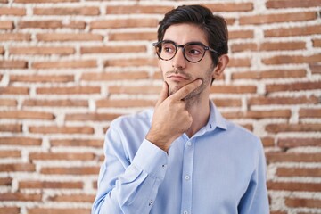 Poster - Young hispanic man standing over brick wall background with hand on chin thinking about question, pensive expression. smiling with thoughtful face. doubt concept.