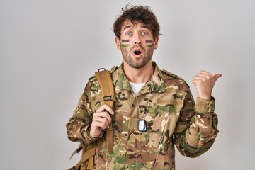 Canvas Print - Hispanic young man wearing camouflage army uniform surprised pointing with hand finger to the side, open mouth amazed expression.