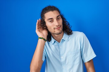 Poster - Young hispanic man standing over blue background smiling with hand over ear listening an hearing to rumor or gossip. deafness concept.