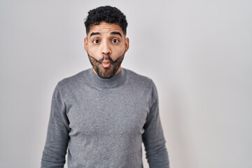 Wall Mural - Hispanic man with beard standing over white background making fish face with lips, crazy and comical gesture. funny expression.