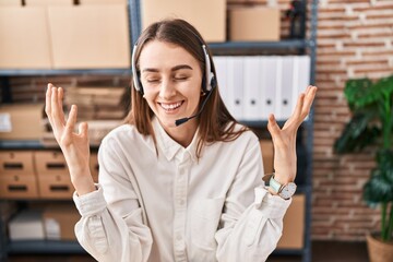 Canvas Print - Young caucasian woman working at small business ecommerce wearing headset celebrating mad and crazy for success with arms raised and closed eyes screaming excited. winner concept