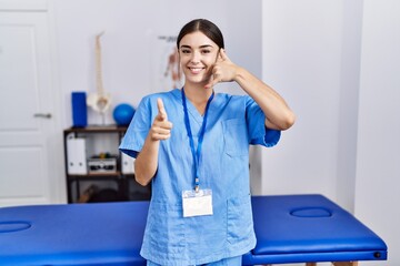 Wall Mural - Young hispanic woman wearing physiotherapist uniform standing at clinic smiling doing talking on the telephone gesture and pointing to you. call me.