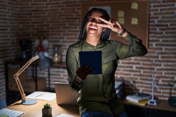 Wall Mural - Young african american with braids working at the office at night doing peace symbol with fingers over face, smiling cheerful showing victory