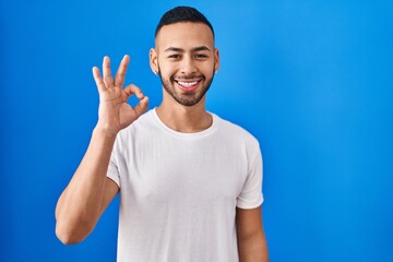 Sticker - Young hispanic man standing over blue background smiling positive doing ok sign with hand and fingers. successful expression.