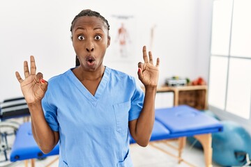 Canvas Print - Black woman with braids working at pain recovery clinic looking surprised and shocked doing ok approval symbol with fingers. crazy expression