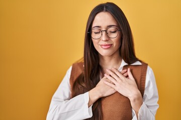 Poster - Young brunette woman standing over yellow background wearing glasses smiling with hands on chest with closed eyes and grateful gesture on face. health concept.