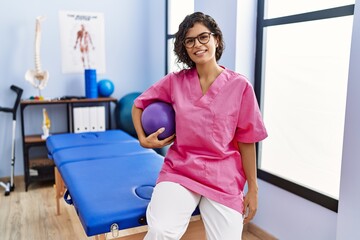 Wall Mural - Young latin woman wearing physiotherapist uniform holding fit ball at clinic