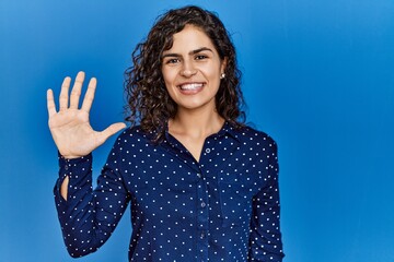 Poster - Young brunette woman with curly hair wearing casual clothes over blue background showing and pointing up with fingers number five while smiling confident and happy.