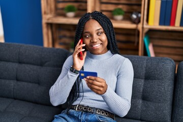 Poster - African american woman talking on smartphone using credit card sitting on sofa at home