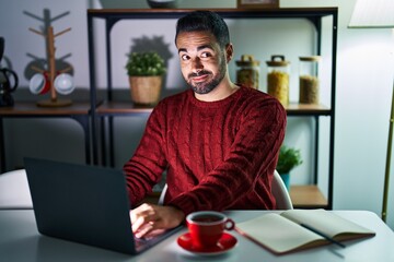 Canvas Print - Young hispanic man with beard using computer laptop at night at home smiling looking to the side and staring away thinking.