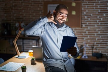 Wall Mural - Young hispanic man with beard and tattoos working at the office at night smiling doing phone gesture with hand and fingers like talking on the telephone. communicating concepts.