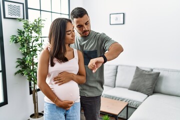 Canvas Print - Young interracial couple expecting a baby, touching pregnant belly checking the time on wrist watch, relaxed and confident