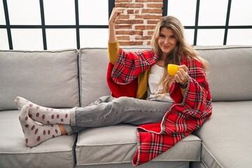 Poster - Young woman sitting on the sofa drinking a coffee at home strong person showing arm muscle, confident and proud of power