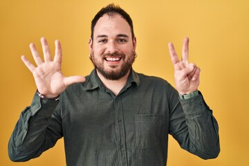 Wall Mural - Plus size hispanic man with beard standing over yellow background showing and pointing up with fingers number seven while smiling confident and happy.